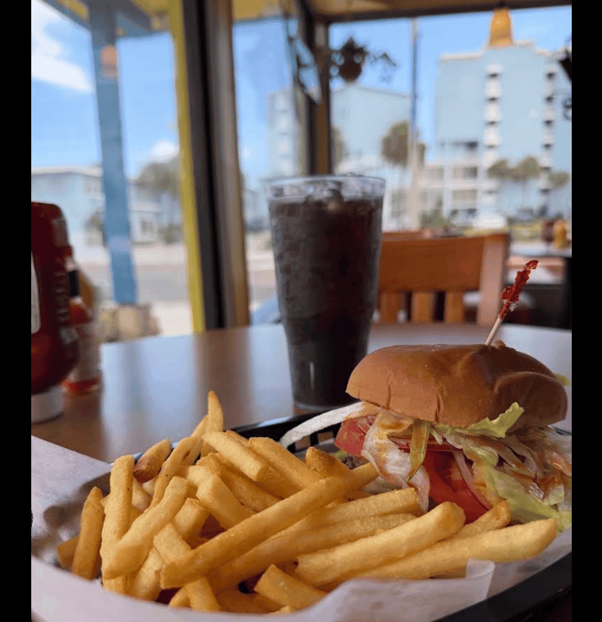 Booth-side view of a burger, fries and drink at J&A Subs & More with the Daytona Beach Shores skyline through the window